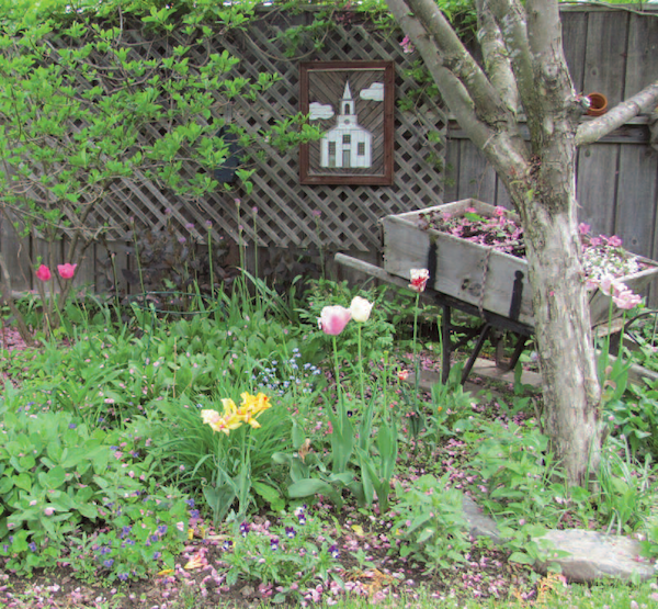 A corner of the garden made more interesting by an old wheelbarrow that has been converted to a container and a picture of a church done in wood.